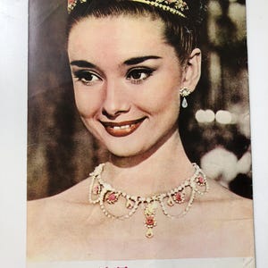 May include: A vintage postcard featuring a black and white photo of Audrey Hepburn in a tiara and necklace. The text "Roman Holiday" is printed in the top right corner. The text "ローマの休日" is printed in the bottom center of the postcard.