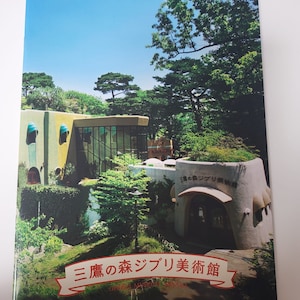 Puede incluir: Portada de libro con el Museo Ghibli en Mitaka, Japón. La imagen muestra un edificio único con un tejado verde, rodeado de exuberante vegetación y un cielo azul claro. El título está en japonés.