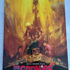 Può includere: Un poster cinematografico vintage de "I Goonies" con una scena del tesoro con un teschio, gioielli e un cappello da pirata. Il poster presenta il titolo del film in rosso e blu, con i nomi di Steven Spielberg e Richard Donner.