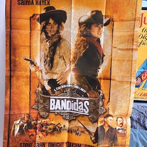 Puede incluir: Un póster de película rectangular de Bandidas con Salma Hayek y Penélope Cruz con sombreros de vaquero y ropa del oeste. El póster incluye el título Bandidas, los nombres del reparto y escenas de acción en la parte inferior en tonos naranjas.