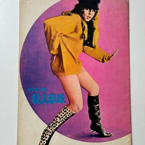May include: Vintage magazine cover with a woman in a mustard yellow sweater, black boots, and a black cap. The background has a purple and pink circular design. The magazine has Japanese text.