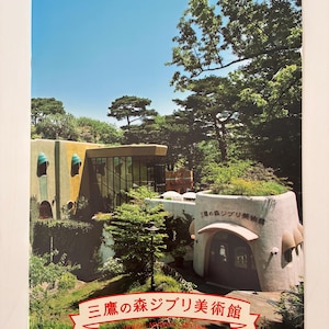 May include: A rectangular brochure featuring a photograph of the Ghibli Museum in Mitaka, Japan. The image displays a multi-level building with green and tan walls, surrounded by lush trees. The text "三鷹の森ジブリ美術館" is printed in red.