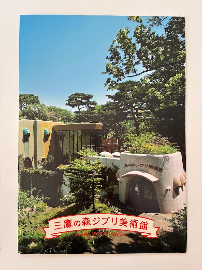 May include: A rectangular brochure featuring a photograph of the Ghibli Museum in Mitaka, Japan. The image displays a multi-level building with green and tan walls, surrounded by lush trees. The text "三鷹の森ジブリ美術館" is printed in red.