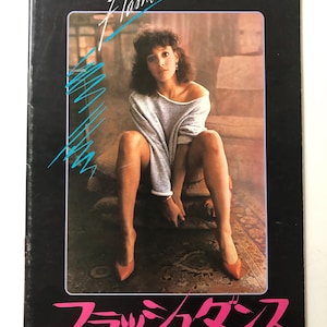 May include: A black movie poster with the title "Flashdance" in a stylized font. The image features a woman in a gray sweater and heels. The Japanese title is also visible in pink.