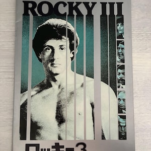 Könnte beinhalten: Ein Filmplakat für "Rocky III" mit einem Porträt von Sylvester Stallone. Das Poster hat einen silbernen Hintergrund mit vertikalen tealfarbenen Streifen. Der Titel "ROCKY III" steht oben, und japanischer Text steht unten.