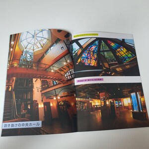 May include: A printed brochure page featuring interior photographs of a building. The left image shows a multi-level hall with a central staircase and the text 吹き抜けの中央ホール. The right images display a stained glass ceiling and a dark room with the text 吹き抜けのフュージングガラス and 常設展示室「動きはじめの部屋」.