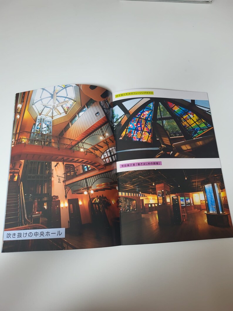 May include: A printed brochure page featuring interior photographs of a building. The left image shows a multi-level hall with a central staircase and the text 吹き抜けの中央ホール. The right images display a stained glass ceiling and a dark room with the text 吹き抜けのフュージングガラス and 常設展示室「動きはじめの部屋」.