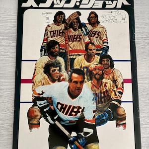 Op de afbeelding: Een vintage filmposter voor "Slap Shot" met een hockeyteam in crème en blauwe shirts met het woord "Chiefs". De poster heeft een zwarte rand en Japanse tekst bovenaan.
