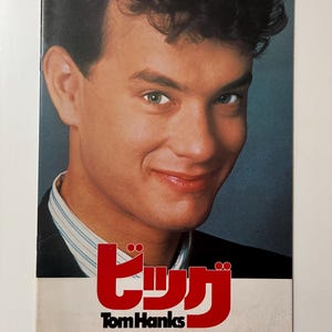 May include: A magazine cover featuring actor Tom Hanks. He is smiling, with green eyes and dark hair. The cover has Japanese text in red and the name "Tom Hanks" in white.