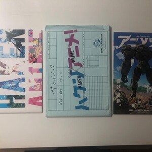 Pode incluir: São exibidos três itens japoneses relacionados a anime. À esquerda, um livro branco com texto japonês e gráficos coloridos. No centro, um envelope azul claro com texto japonês. À direita, uma revista com uma ilustração de robô e texto japonês.