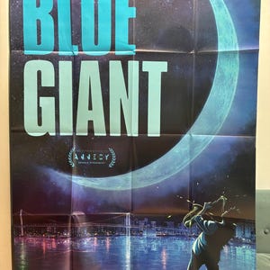 Puede incluir: Un póster de "BLUE GIANT" presenta texto turquesa grande sobre un fondo oscuro y estrellado. También se ven una luna creciente y una figura silueteada tocando el saxofón. El póster incluye el texto "ANNECY SEANCE EVENEMENT".