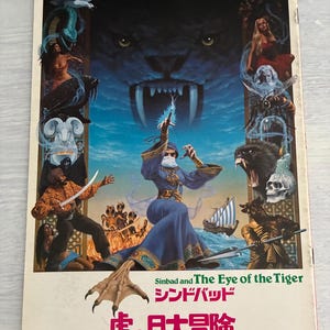 May include: A vintage book cover for 'Sinbad and The Eye of the Tiger'. The illustration shows a fantasy scene with a tiger's face, a sorceress, and mythical creatures. The title is in English and Japanese.