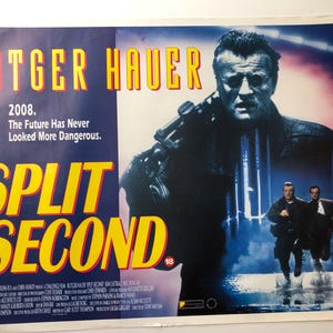 Może przedstawiać: Plakat filmowy do filmu "Split Second" z Rutgerem Hauerem. Tytuł jest dużymi żółtymi literami, z nazwiskiem aktora powyżej. Plakat ma ciemnoniebieskie tło ze zdjęciami aktora i innych postaci.