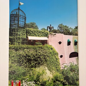 May include: A rectangular print featuring a pink building with green accents, a tall metal cage-like tower covered in ivy, and a dark robot-like figure on the roof. The scene includes lush green foliage, white flowering bushes, and a clear blue sky.