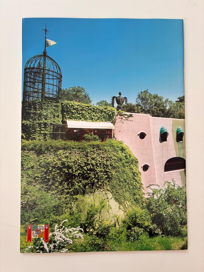 May include: A rectangular print featuring a pink building with green accents, a tall metal cage-like tower covered in ivy, and a dark robot-like figure on the roof. The scene includes lush green foliage, white flowering bushes, and a clear blue sky.