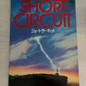 Peut inclure: Affiche du film "Short Circuit" avec un ciel dramatique. Le titre est en rouge, avec du texte japonais en dessous. Un robot en silhouette se tient sur une colline, avec un éclair descendant des nuages.
