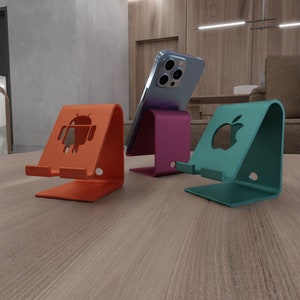May include: Three phone stands made of metal. The stand on the left is orange and has an Android logo. The stand in the middle is purple and has a phone on it. The stand on the right is teal and has an Apple logo.