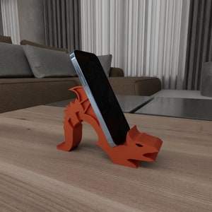 May include: A 3D printed orange dragon phone stand with a black smartphone resting in the stand. The stand is on a wooden table.