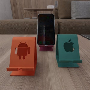 Op de afbeelding: Drie telefoonstandaards van metaal met verschillende logo's. De standaard links is oranje en heeft een Android-logo. De standaard in het midden is roze en heeft geen logo. De standaard rechts is groen en heeft een Apple-logo.