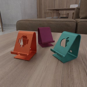 Op de afbeelding: Drie telefoonstandaards van metaal, elk met een ander uitgesneden logo. De standaard links heeft een Android-logo, de middelste standaard is paars en de standaard rechts heeft een Apple-logo. De standaards staan op een houten oppervlak.