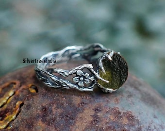 Puede incluir: Un anillo de plata con una banda diseñada para parecer ramas de árbol retorcidas, con un pequeño detalle de flor y una piedra circular texturizada de color verde oliva oscuro. El texto Silvertreesindia es visible en el lateral.