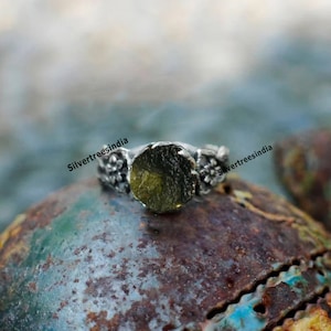 K&ouml;nnte beinhalten: Ein silberner Ring mit einem runden, gr&uuml;nen Stein, der in der Mitte eingesetzt ist. Die Band ist mit kleinen floralen Designs verziert. Der Ring liegt auf einer rostigen Oberfl&auml;che.