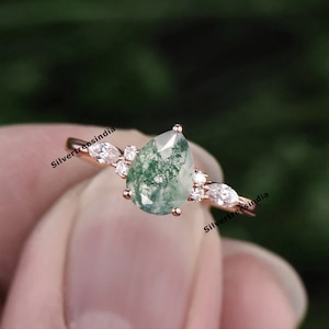 May include: A pear-shaped green moss agate ring set in rose gold with a band of smaller clear stones. The ring is on a finger.