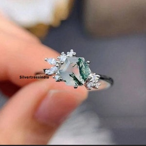 Puede incluir: Un anillo de plata con una piedra preciosa hexagonal de ágata verde musgo rodeada de pequeñas piedras preciosas transparentes. El anillo se lleva en un dedo.