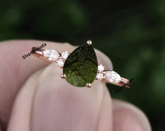 May include: Rose gold ring featuring a pear-shaped, dark green gemstone as the centerpiece. The ring is accented with small, round, and marquise-cut white gemstones. The ring's band is thin and delicate, with a polished finish.
