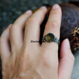 K&ouml;nnte beinhalten: Ein silberner Ring mit einem gr&uuml;nen Edelstein, der in eine strukturierte Band gefasst ist. Der Ring wird an einem Finger getragen.