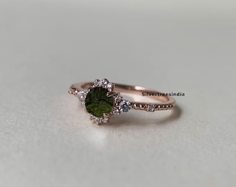 May include: A rose gold ring with a green gemstone surrounded by small clear stones. The ring has a delicate design with a textured band. The text "Silvertreeindia" is visible on the band.