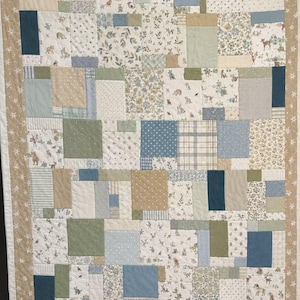 May include: A patchwork quilt featuring a variety of rectangular fabric blocks in shades of blue, sage green, beige, and white. The design includes patterns of small woodland animals, floral motifs, stripes, polka dots, and plaid, finished with a tan border.