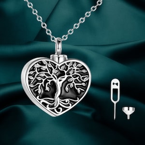 Może przedstawiać: Srebrny naszyjnik z zawieszką w kształcie serca z motywem drzewa życia. Zawieszka ma czarne tło i szczegółową ilustrację drzewa. Zawiera mały lejek i narzędzie do napełniania. Naszyjnik jest prezentowany na tle w kolorze morskim.