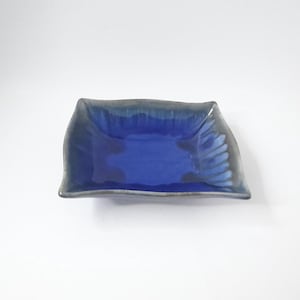 May include: A small, square ceramic dish with a blue glaze and a slightly wavy rim. The dish has a light blue interior and a darker blue exterior.