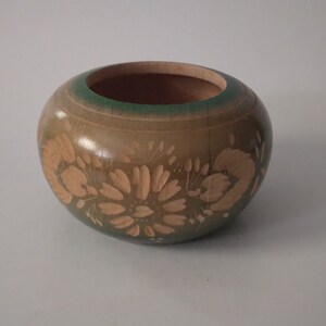 May include: A small, round wooden bowl with a green rim and a floral design carved into the surface. The bowl is painted in shades of brown and green.