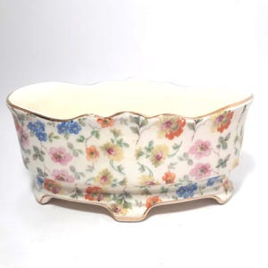 Op de afbeelding: Een vintage porseleinen bloempot met een crèmekleurige binnenkant en een geschulpte rand. De buitenkant is versierd met een bloemmotief in roze, geel, oranje en blauw op een witte achtergrond. De pot heeft een gouden rand en een voet.