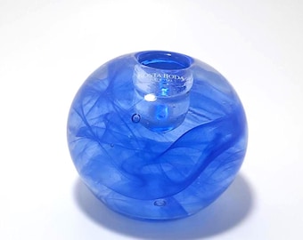 May include: A spherical, blue glass candle holder with a smaller, cylindrical holder on top. The glass has a swirled pattern and the text "KOSTA BODA" is visible on the top. A decorative item for home decor.