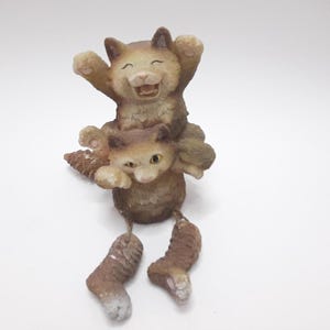 以下が含まれることがあります： 茶色と白のセラミック製の猫の置物2体。上の猫は腕を上げて口を開けて笑っている。下の猫は足をぶら下げて頭を横に傾けている。