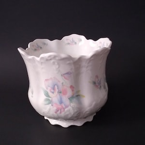 May include: White ceramic planter with a scalloped rim and a bulbous body. The planter is decorated with a floral design featuring pink, purple, and blue flowers. The planter has a textured surface and a decorative base.