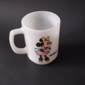 May include: White ceramic mug with a cartoon image of Minnie Mouse wearing a pink dress and yellow bow. The mug has a black handle and the word "Minnie" is printed on the bottom.