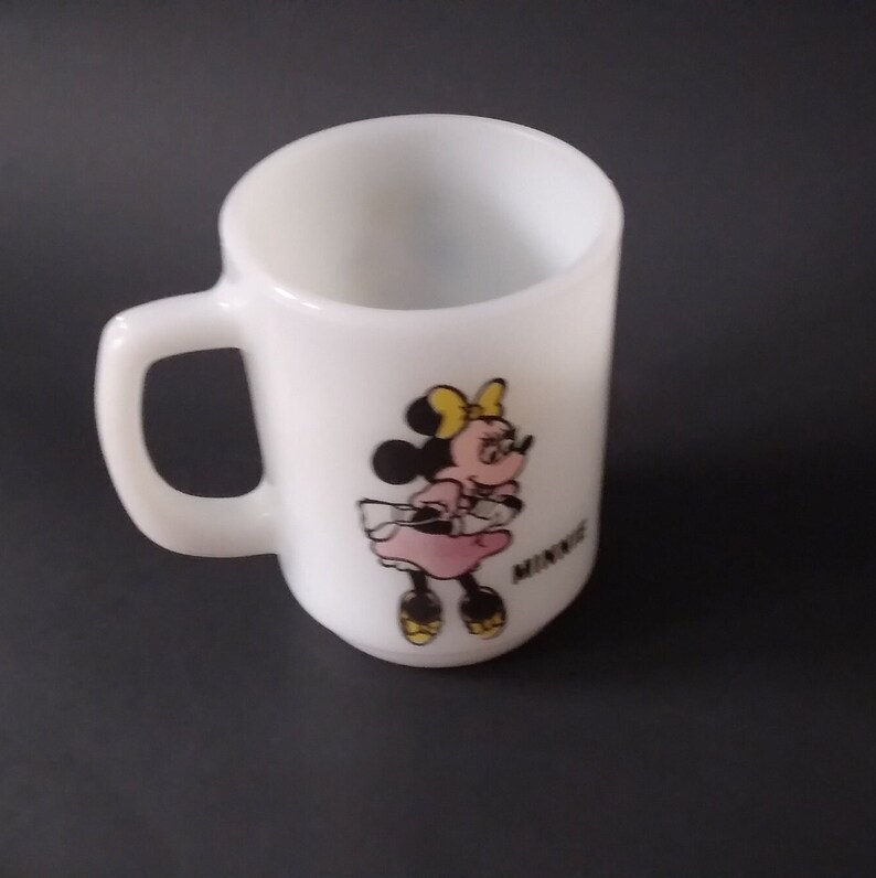 May include: White ceramic mug with a cartoon image of Minnie Mouse wearing a pink dress and yellow bow. The mug has a black handle and the word "Minnie" is printed on the bottom.