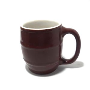 May include: A brown ceramic mug with a white interior and a rounded handle. The mug has a barrel-like design with horizontal ridges. The mug is set against a white background.