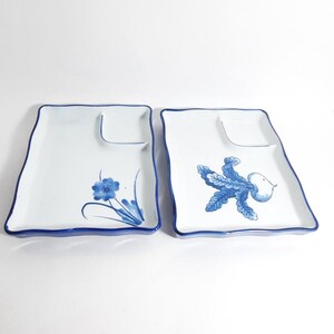 Puede incluir: Dos platos de servir de cerámica blanca con borde azul y diseños florales. Un plato presenta una flor azul con hojas verdes, mientras que el otro tiene un diseño de hojas azules con una fruta blanca.