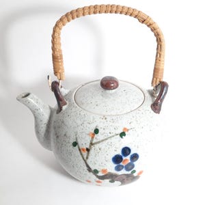 Peut inclure: Une théière en céramique blanche avec une poignée brune et une poignée de couvercle en bambou tissé. La théière a un motif floral avec des accents bleus, orange et verts.