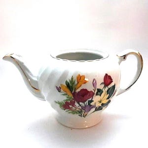 May include: White teapot with a floral design featuring red, yellow, and purple flowers. The teapot has a gold trim around the lid and spout.