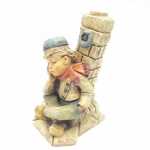 以下が含まれることがあります： 青い帽子と赤いマフラーをつけた少年の小さな陶器の置物で、石造りの構造物に座っています。少年は頭を手に乗せて眠っているようです。