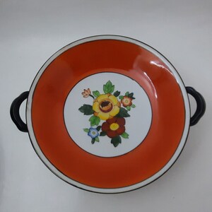 Puede incluir: Un plato de servir de cerámica blanca con dos asas negras. El plato tiene un borde ancho de color naranja con una línea negra alrededor del borde. El centro del plato presenta un círculo blanco con un diseño floral en tonos de amarillo, rojo, naranja, azul y verde.