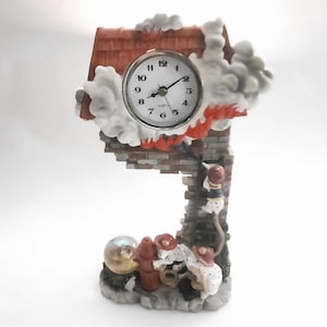 Puede incluir: Un reloj de cerámica con forma de casa con un techo rojo y humo saliendo de la chimenea. La esfera del reloj es blanca con números y manecillas negras. Hay figuritas de bomberos en el lateral de la casa.