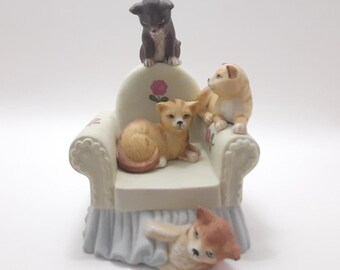 Puede incluir: Una figurita de porcelana de cuatro gatos en un sillón blanco. Los gatos están en varias poses, con un gato sentado en el respaldo del sillón, un gato sentado en el brazo del sillón, un gato acostado en el asiento del sillón y un gato asomándose desde debajo del sillón.