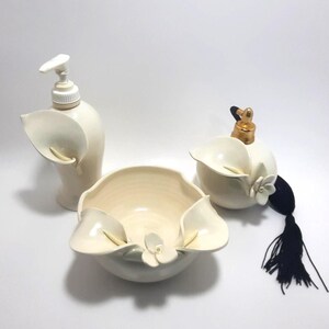May include: A three-piece ceramic bathroom set in off-white featuring a soap dispenser with a pump, a small bowl, and a perfume atomizer with a gold-tone nozzle and a black tassel. Each piece is decorated with sculpted calla lily flower motifs.